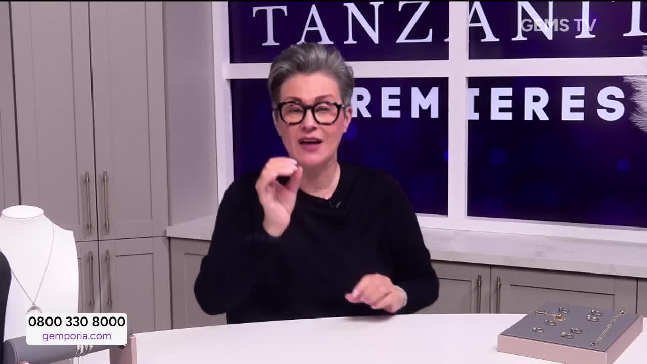 The woman on Gems TV in the United Kingdom gestures with her hands as she speaks. Behind her, the words "Tanzanite Premieres" are displayed on a screen.
