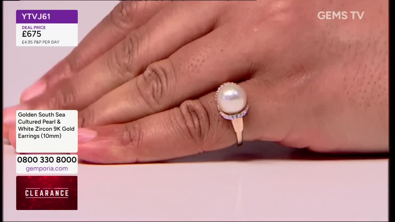 A hand, with a ring featuring a large, round pearl, is held up for display. The ring's band is gold, and the pearl is surrounded by a halo of smaller stones.

