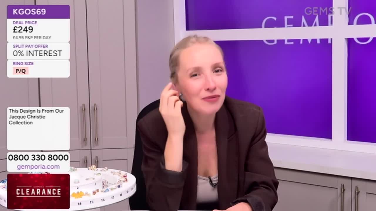 A woman in a brown blazer touches her ear, likely adjusting an earring, while looking toward the camera. Beside her, a screen displays the price of a ring and the Gems TV logo.
