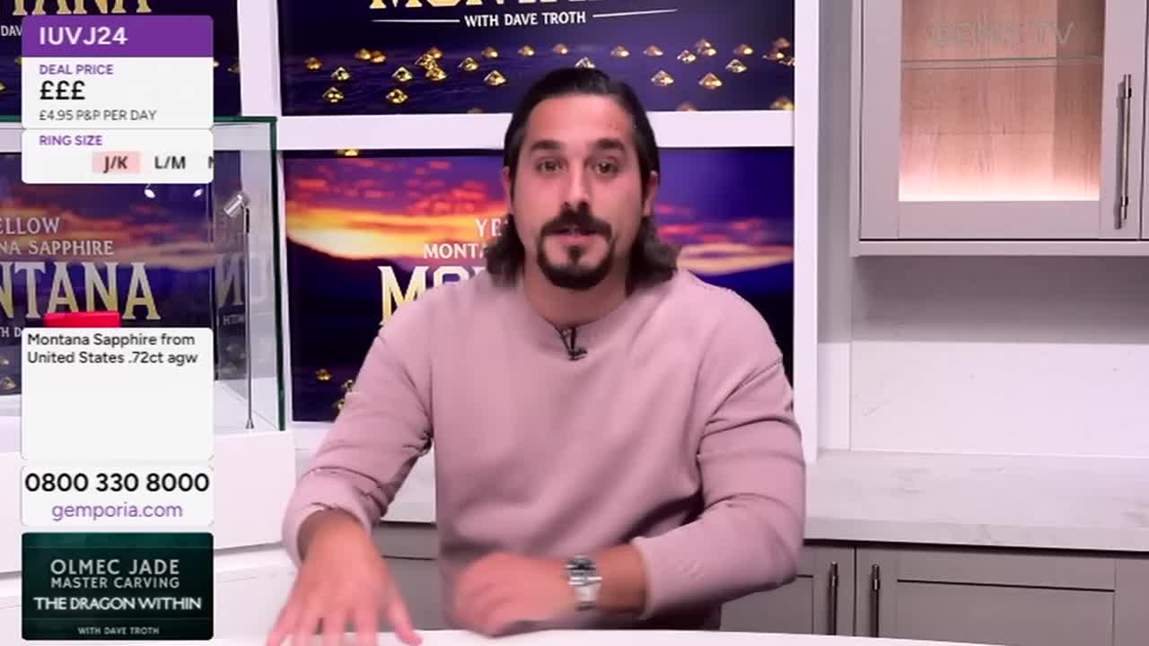 A man in a pink sweater gestures with his hands, speaking directly to the camera. Behind him, a display case shows a gem, and graphics with prices and ring sizes are visible.
