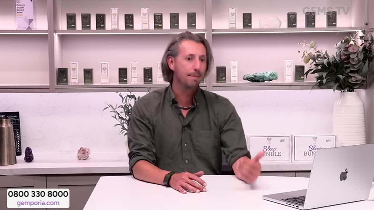The man in the green shirt is gesturing towards the laptop, likely explaining something. Behind him, shelves display various products, and the Gems TV logo is visible in the corner.

