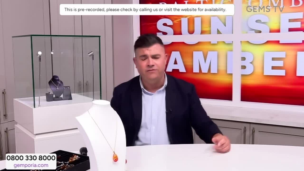 A man in a blazer sits at a desk, speaking directly to the camera. Behind him, the "Sunset Amber" segment of Gems TV is displayed, along with jewelry on display.
