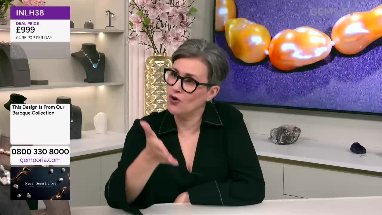 A woman wearing glasses gestures emphatically, her hand pointing towards the screen. Behind her, a necklace of large, orange pearls is displayed on Gems TV.
