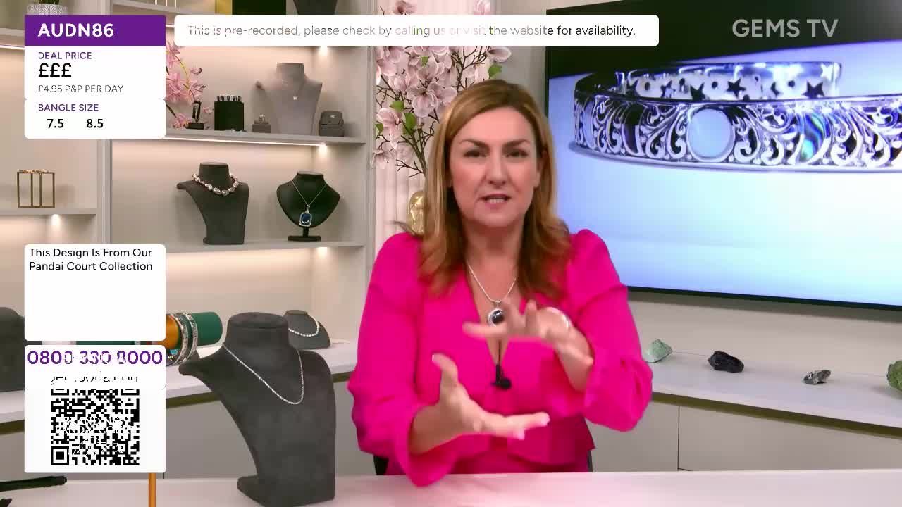 A woman in a bright pink top gestures with her hands, describing jewelry on a screen behind her.  The Gems TV presenter is surrounded by displays of necklaces and bangles.

