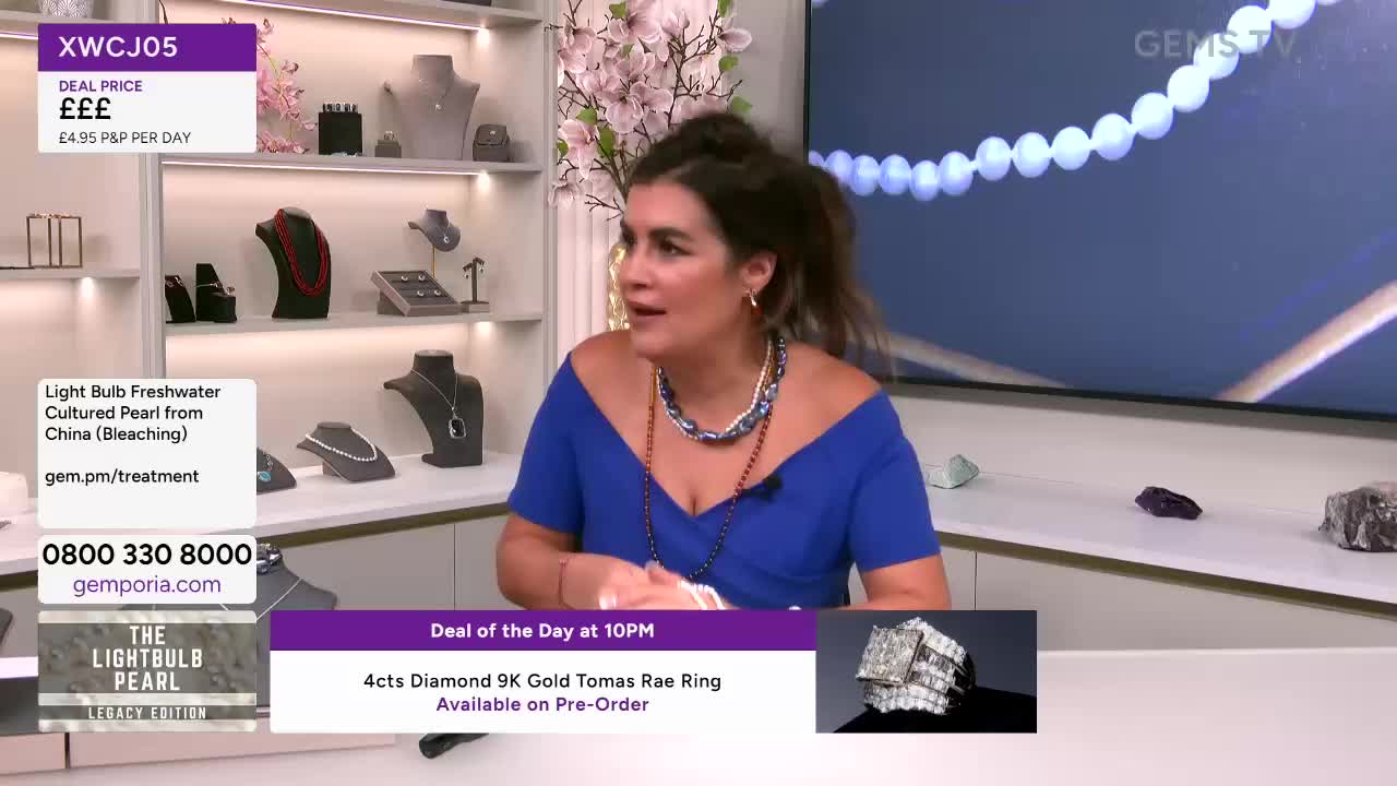 A woman in a blue dress gestures animatedly, speaking to someone off-camera. Behind her, a display of jewelry sits on shelves, and a Gems TV logo is visible in the corner.

