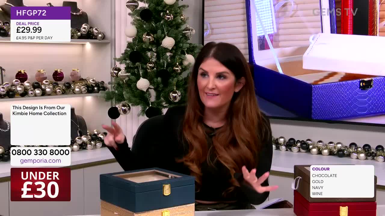 The woman on Gems TV gestures towards a stack of jewelry boxes, while a larger open one is displayed on a screen behind her. The boxes are in various colors, with the price of a featured item displayed on the screen.
The woman on Gems TV gestures towards a stack of jewelry boxes, while a larger open one is displayed on a screen behind her. The boxes are in various colors, with the price of a featured item displayed on the screen.