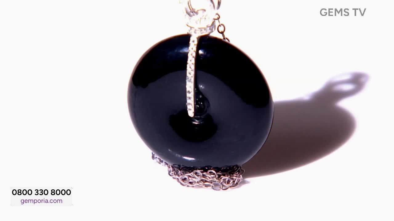 On Gems TV, a large, glossy black pendant hangs suspended against a bright white background. A silver chain and a diamond-studded bale support the round gemstone, which casts a long shadow.
On Gems TV, a large, glossy black pendant hangs suspended against a bright white background. A silver chain and a diamond-studded bale support the round gemstone, which casts a long shadow.