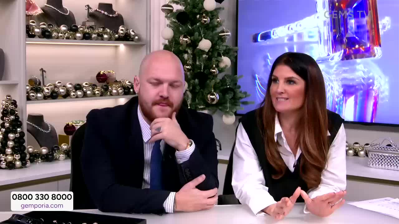 The woman gestures with her hands as she speaks, while the man next to her rests his chin on his hand. Behind them, a screen displays a close-up of a large gemstone, and a Christmas tree is visible.
The woman gestures with her hands as she speaks, while the man next to her rests his chin on his hand. Behind them, a screen displays a close-up of a large gemstone, and a Christmas tree is visible.