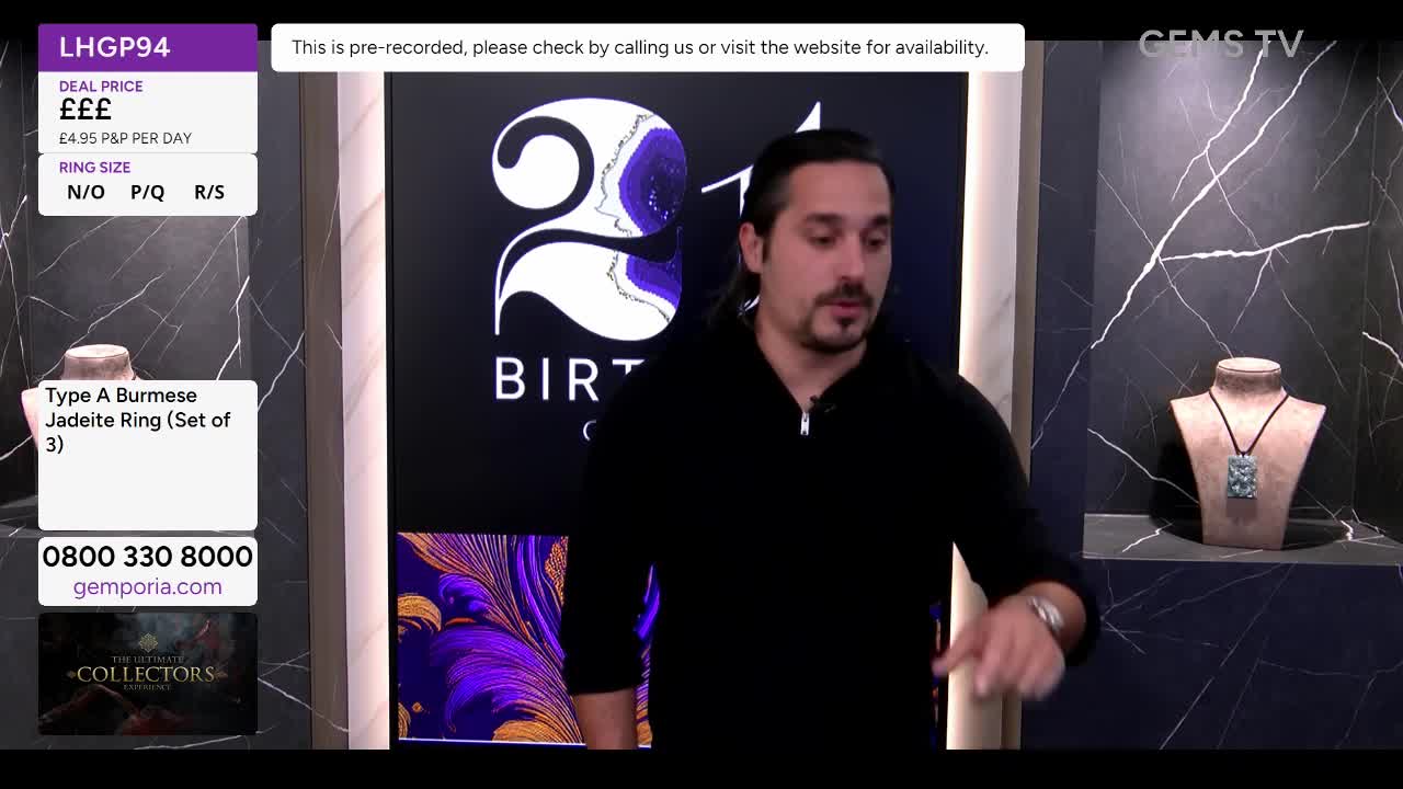 A man in a black sweater gestures towards the jewelry displays on Gems TV. The screen shows a pre-recorded segment, with details about a Burmese jadeite ring set.
A man in a black sweater gestures towards the jewelry displays on Gems TV. The screen shows a pre-recorded segment, with details about a Burmese jadeite ring set.