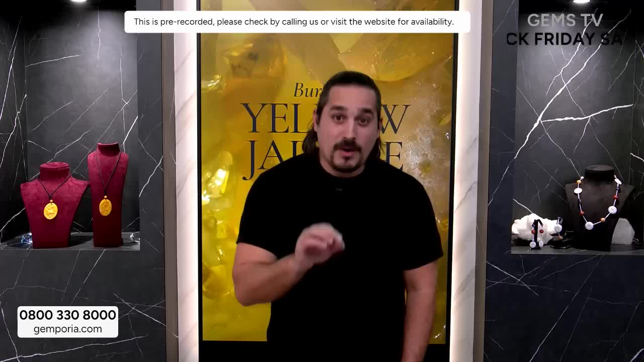 A man in a black shirt gestures emphatically while speaking, likely on Gems TV in the United Kingdom. Behind him, jewelry displays sit on either side, with a large "Yellow Jade" sign in the background.
