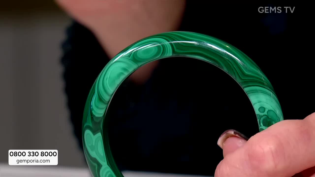 A hand holds up a polished malachite bangle, showcasing its swirling green patterns. The GEMS TV logo is visible in the corner, and a phone number is displayed at the bottom.
