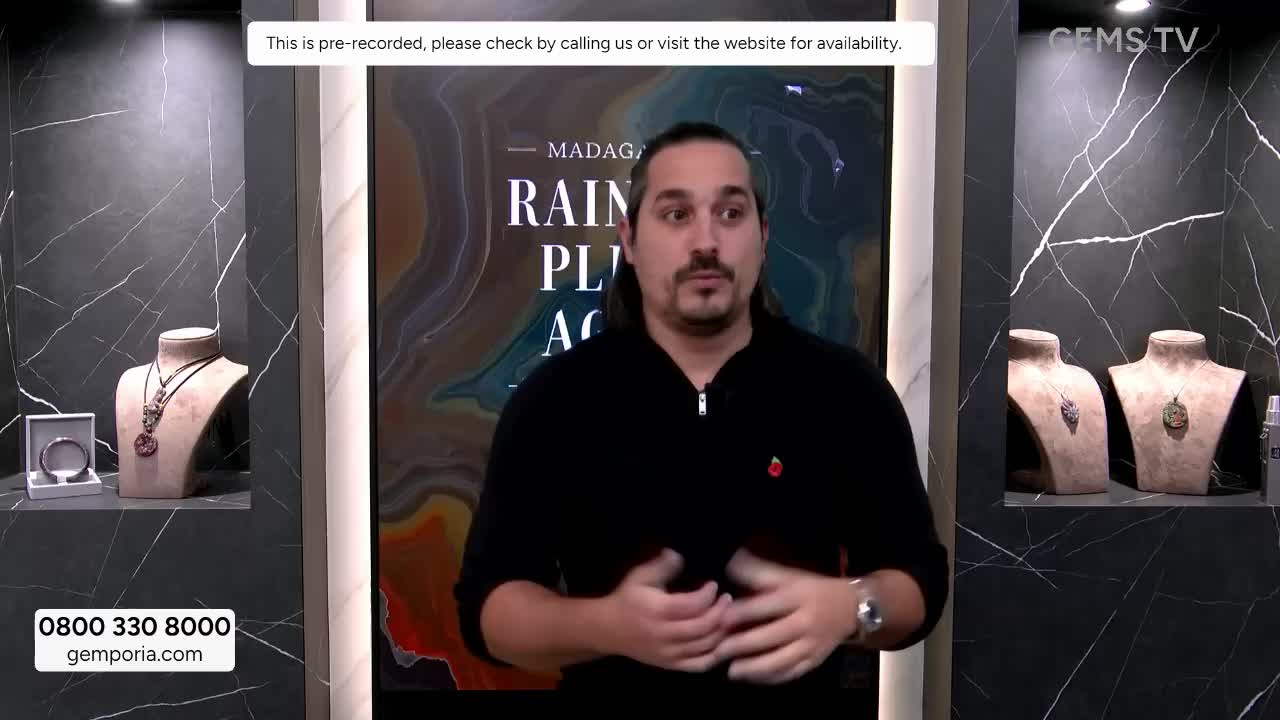 A man in a black sweater gestures with his hands, speaking directly to the camera on a Gems TV set. Jewelry displays are on either side of him, and the broadcast is pre-recorded.
