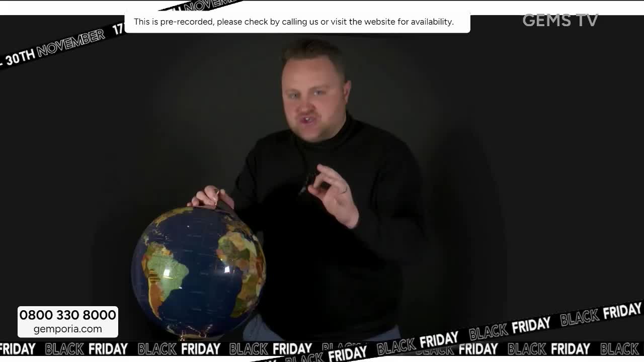 A man in a black sweater gestures with his right hand as he holds a globe. The Gems TV broadcast is pre-recorded, and it's Black Friday.
