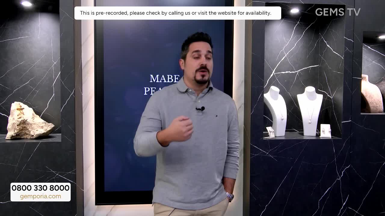 A man in a grey shirt gestures with his right hand as he speaks, a microphone clipped to his collar. Behind him, on Gems TV, are displays of jewelry and a large screen with text.
