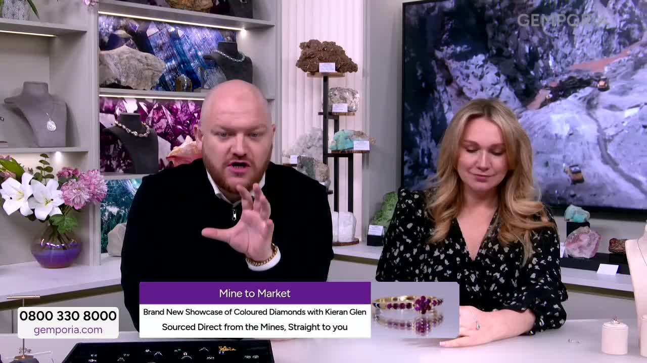 A man gestures emphatically while speaking, a woman beside him looks down at a bracelet. The Gems TV screen behind them displays a vast open-pit mine.