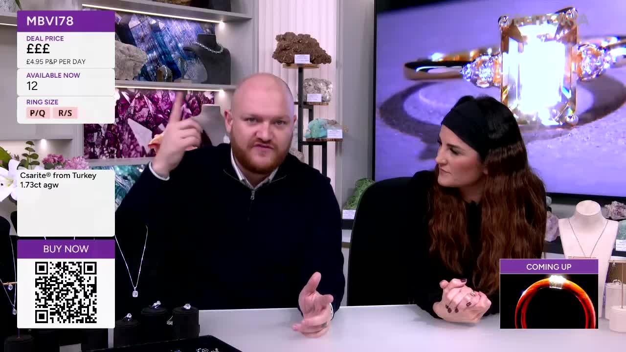 A man gestures emphatically while a woman listens intently on Gems TV. Behind them, a large screen displays a sparkling ring, and shelves hold colourful crystals.