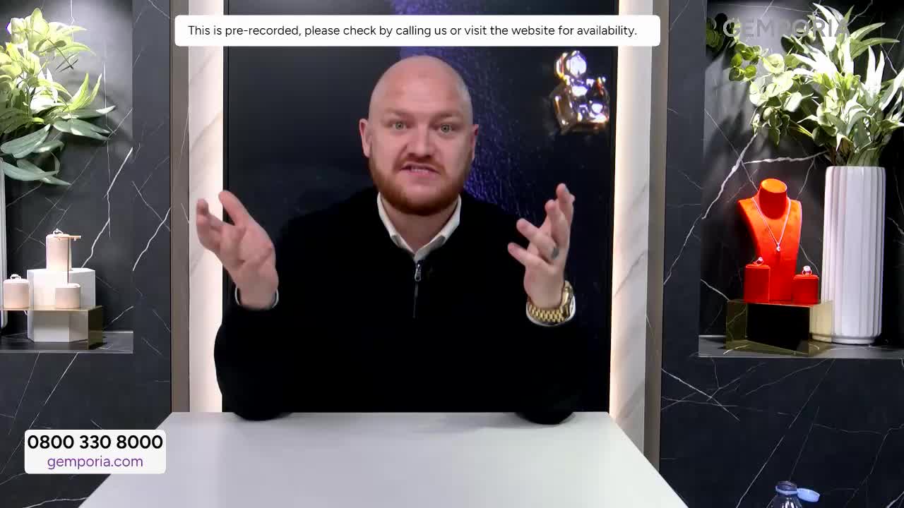 A man with a bald head and beard gestures with both hands as he speaks. On a shelf to his right, a red bust displays a silver necklace.
