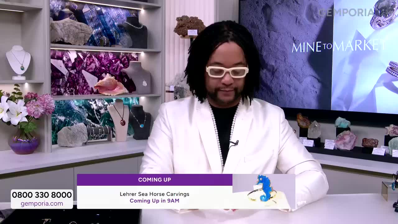 A presenter in a white suit stands before shelves of gemstones and jewelry. A screen behind him displays "Mine to Market" and a preview of Lehrer Sea Horse Carvings coming up at 9 AM.