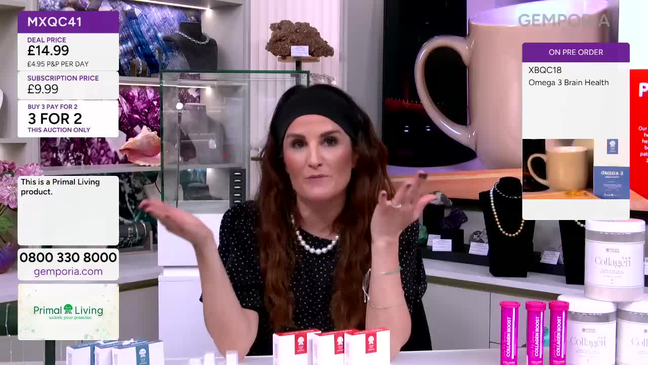A woman gestures with both hands while speaking, her dark hair pulled back by a black headband. On the table before her sit three small white boxes, and behind her, a large white mug is displayed.
