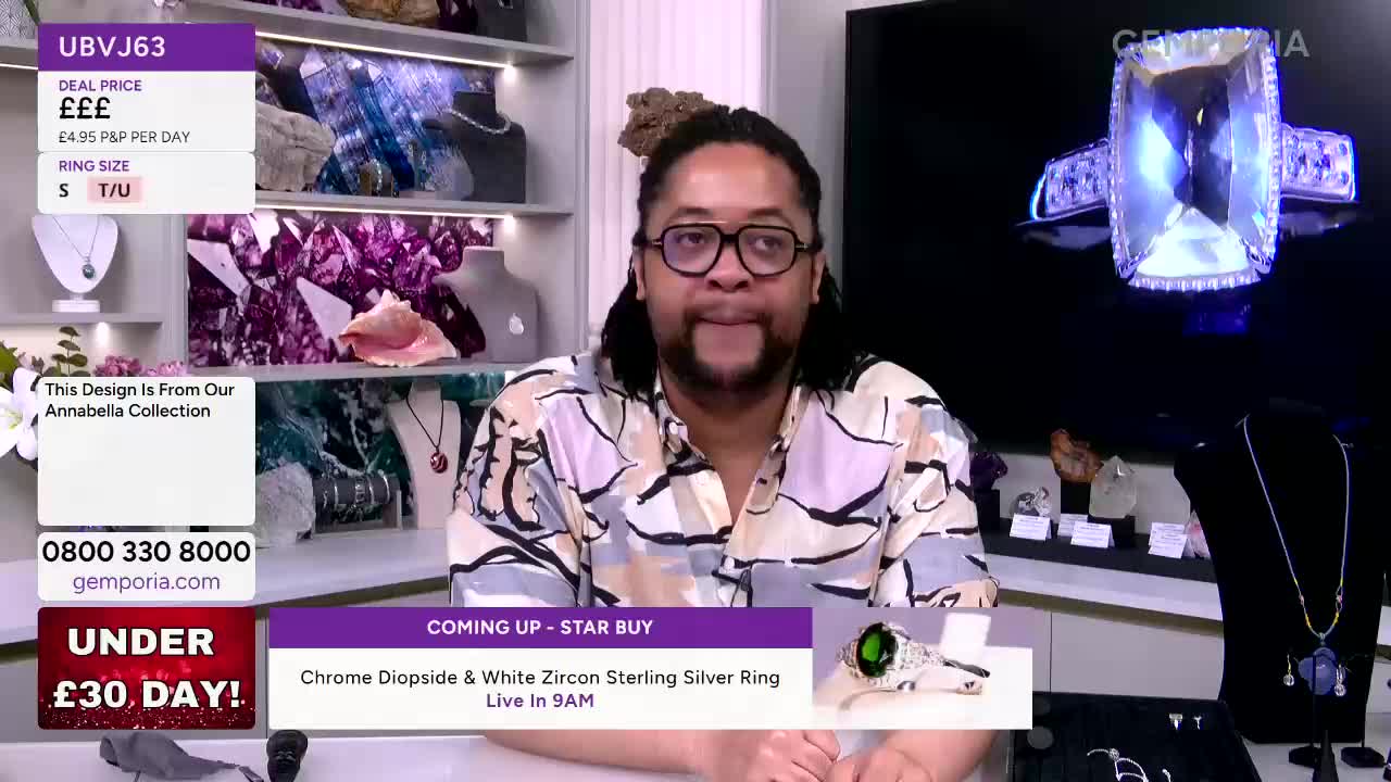 A man in a patterned shirt stands in front of shelves displaying jewellery. A large, sparkling ring is shown on a screen behind him.