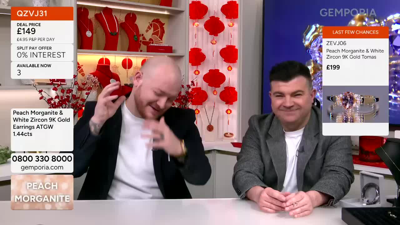 The man on the left is holding up a small red box, tilting his head back with a grin. Beside him, the other man smiles, his hands resting on the table.