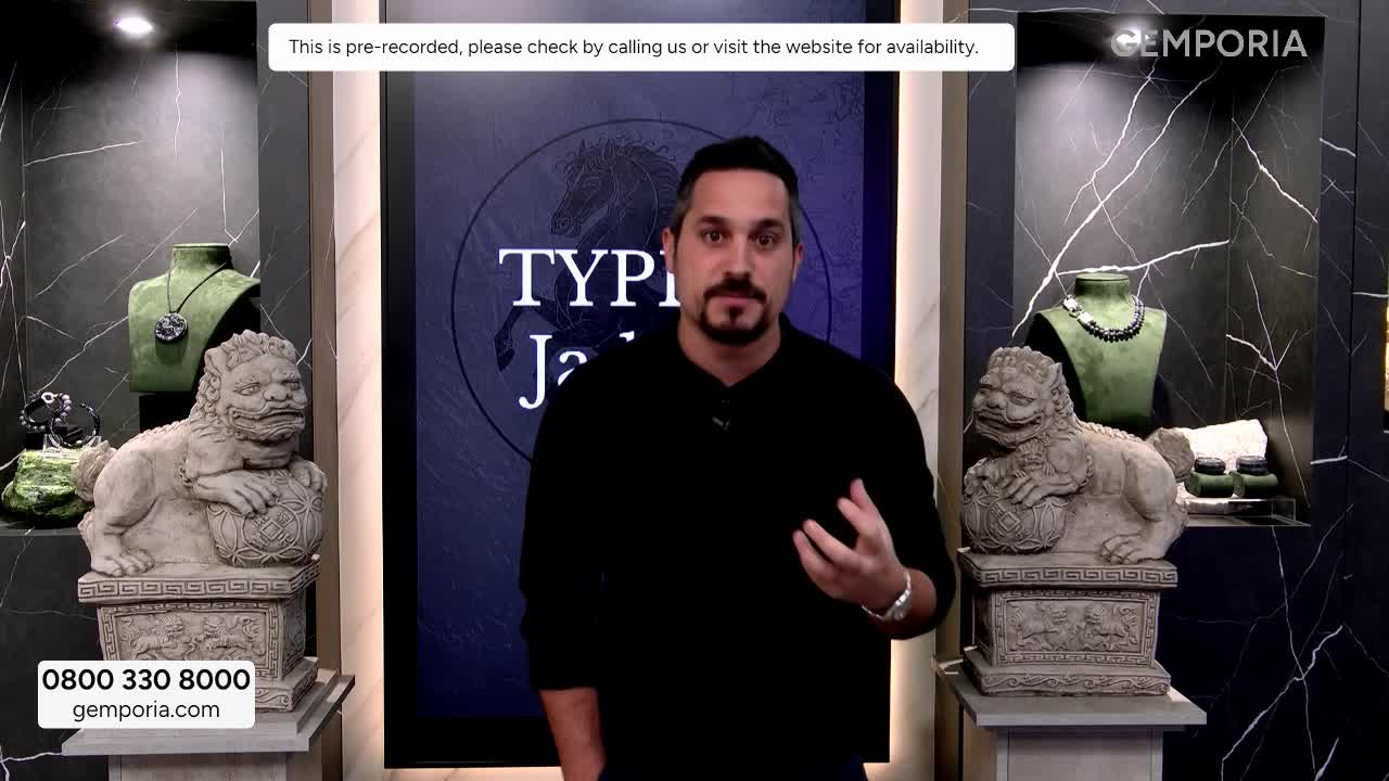 A man in a black shirt gestures with his hands while speaking in front of a display of jade jewelry. The Gems TV logo is visible in the background.