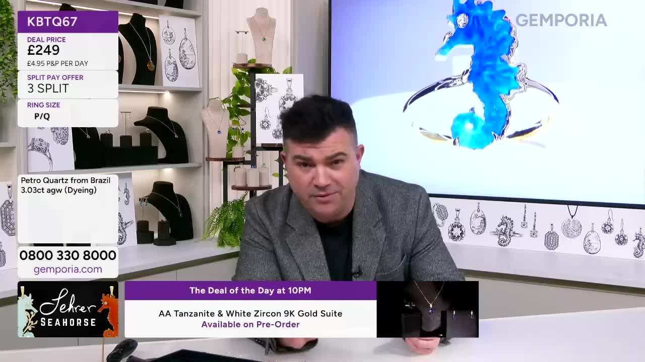 A man in a grey jacket is speaking directly to me. Behind him, a large screen displays a bright blue seahorse pendant, and shelves are filled with necklaces on display.