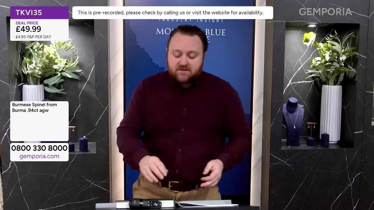 A man in a maroon shirt gestures with his hands as he talks. On a shelf to his right, a necklace rests on a blue bust, flanked by a tall white vase of greenery and a small ring stand.