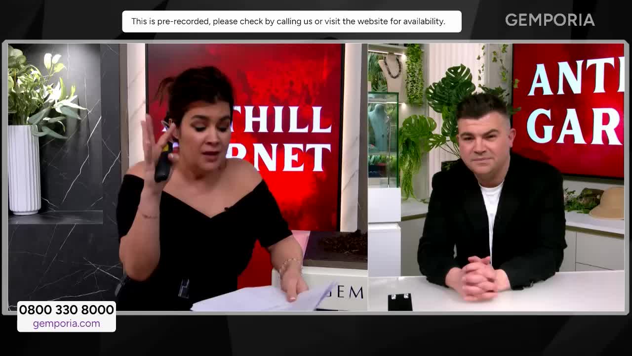 A woman in a black off-the-shoulder top gestures with a remote in her hand while speaking. Beside her, a man in a black jacket sits with his hands clasped, looking towards the camera.