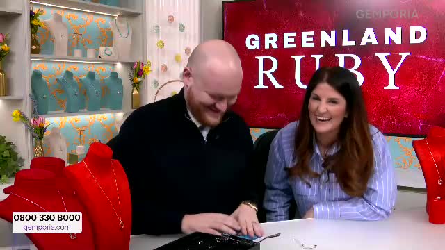 A man and woman are laughing heartily on a television set for Gems TV. Behind them, a large screen displays "GREENLAND RUBY."