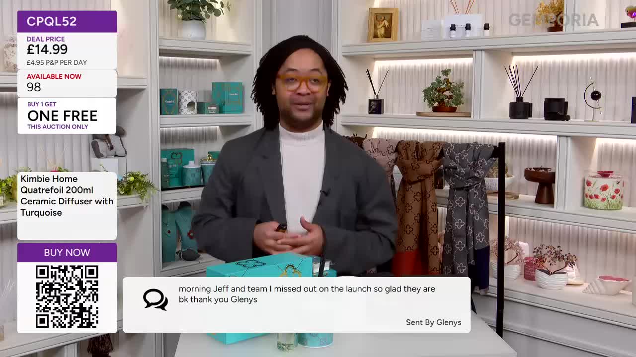A man in a grey blazer stands before shelves stocked with diffusers and decorative items. Scarves with a repeating pattern hang on a rack to his right.