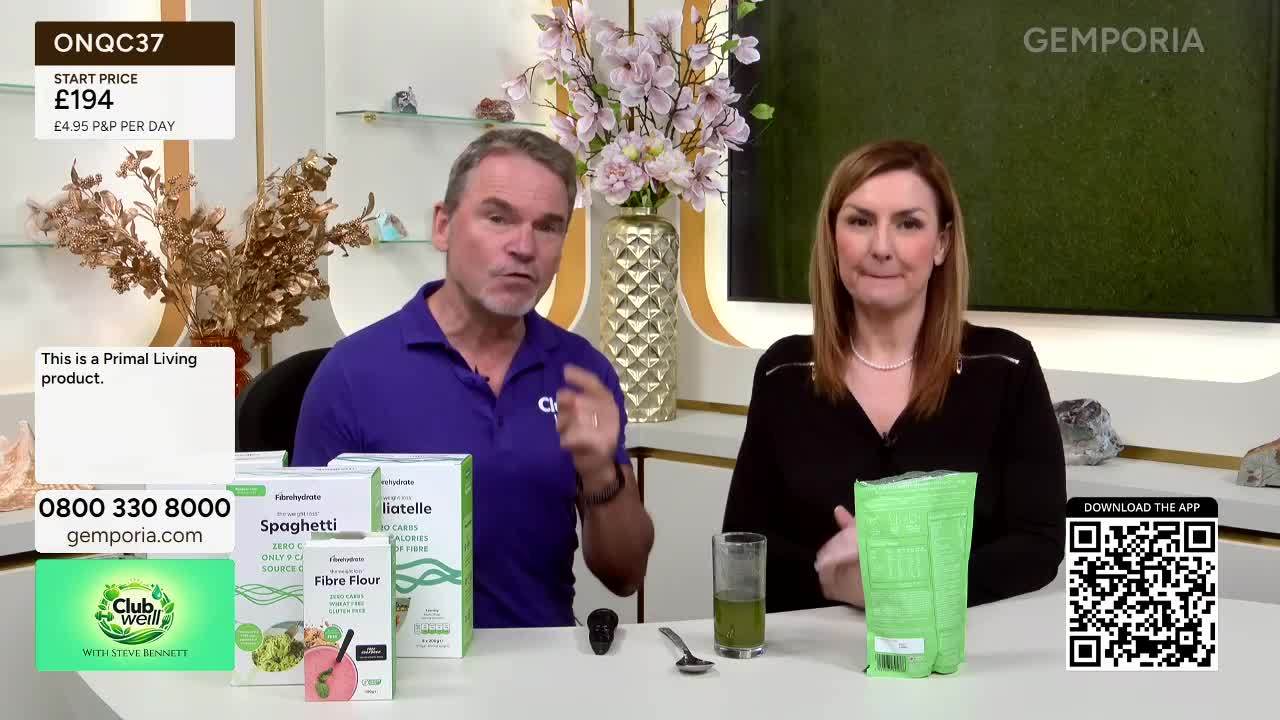 A man in a purple shirt gestures emphatically while a woman in black stands beside him, holding a green packet. Boxes of what appears to be pasta and fibre flour are arranged on the table between them.