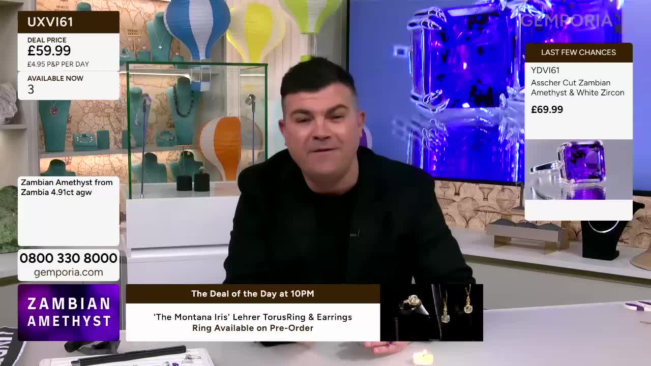 A presenter is talking live on Gemporia, with jewellery displays behind him. A large screen shows a close-up of a faceted amethyst ring.