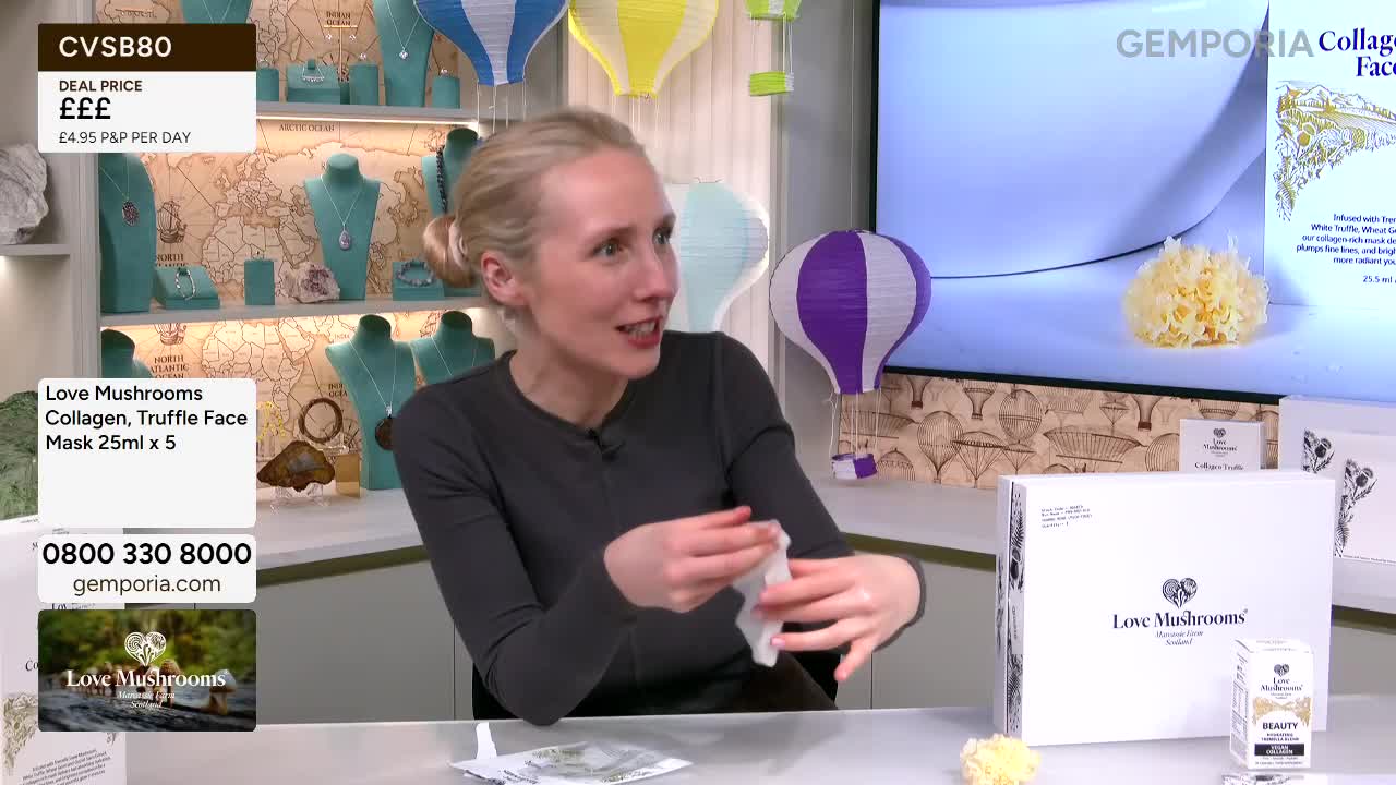 A woman on a Gems TV set is holding a small white wipe. Behind her, shelves display jewellery, and colourful paper lanterns hang in the air.