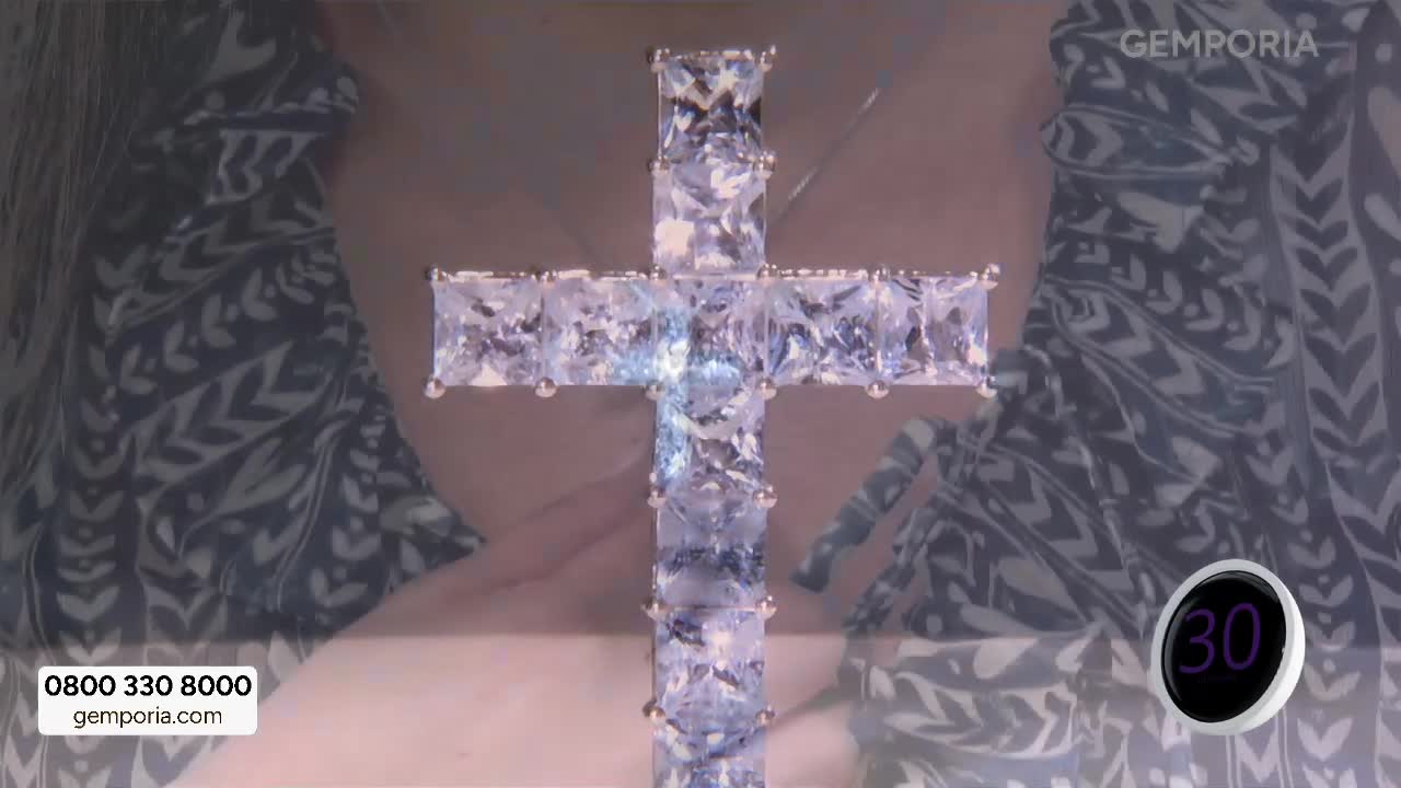 A large, sparkling cross made of clear, rectangular stones hangs centrally. The presenter's patterned dress, a common sight on Gems TV, is visible behind it.