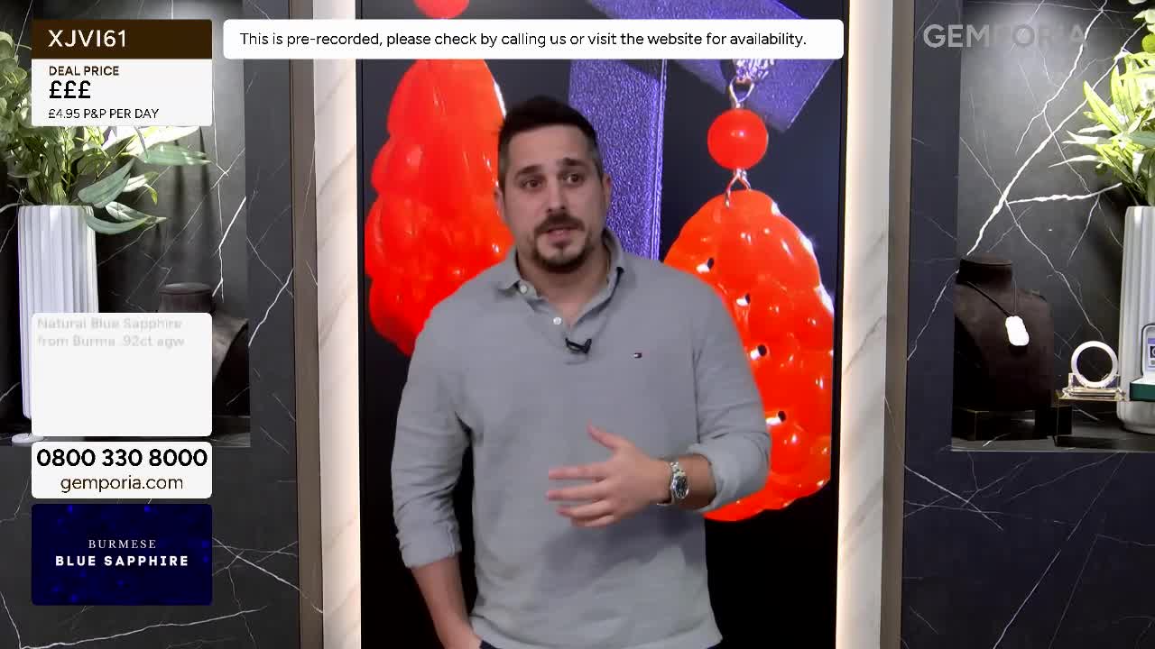 A man in a grey polo shirt gestures as he speaks. Behind him, large, vibrant orange pendant earrings hang against a dark background. A man in a grey polo shirt gestures as he speaks. Behind him, large, vibrant orange pendant earrings hang against a dark background.