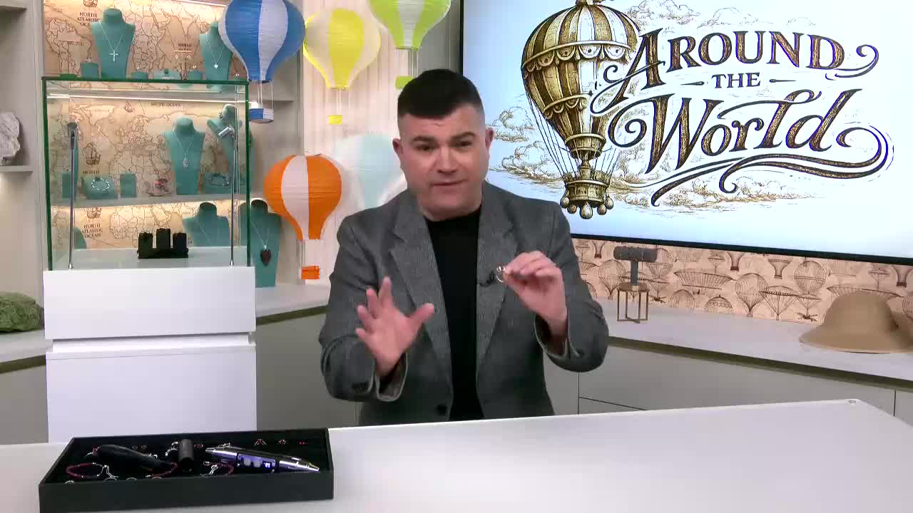 A man in a grey jacket gestures with his hands, holding up a ring for the camera. Behind him, a screen displays "Around the World" with a hot air balloon graphic.