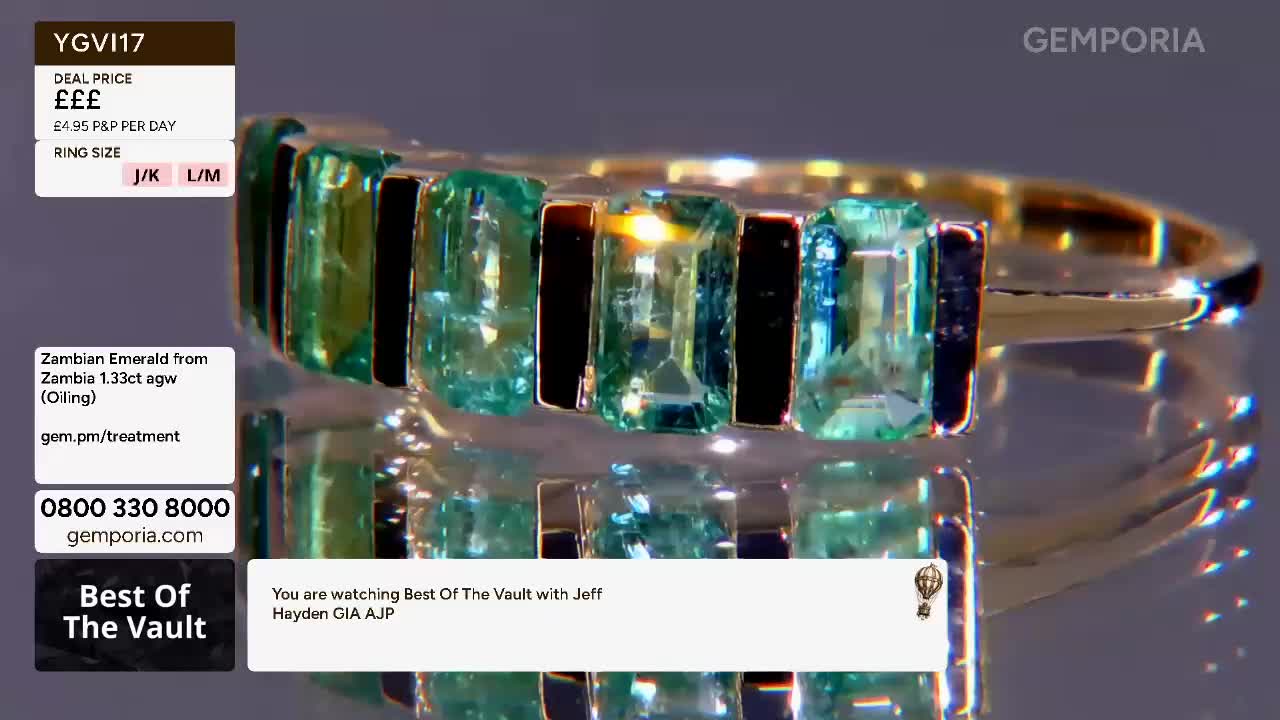 A gold ring with emeralds and black stones sits on a reflective surface. The gems catch the light, showing their facets and colour.