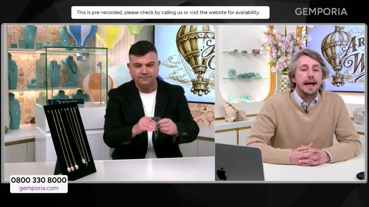 A man in a black jacket holds up a delicate necklace, examining it under the studio lights. Beside him, another presenter speaks animatedly, his hands clasped on the desk in front of him.