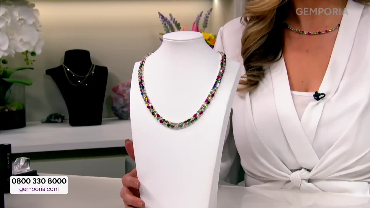 A woman in a white blouse presents a vibrant gemstone necklace on a display stand. Another necklace, equally colourful, rests on her neck, catching the light.