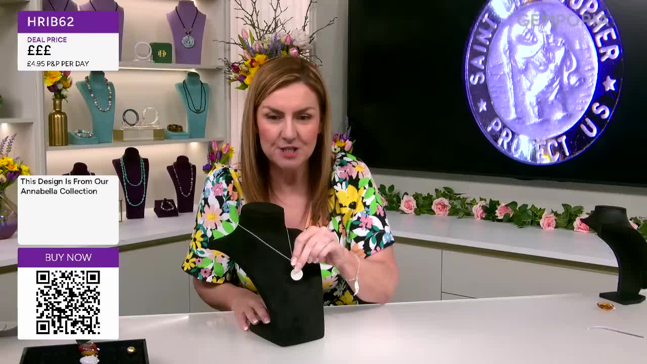 The presenter holds up a delicate silver necklace, its pendant catching the light. Behind her, a screen displays a Saint Christopher medal, a common motif for travellers.