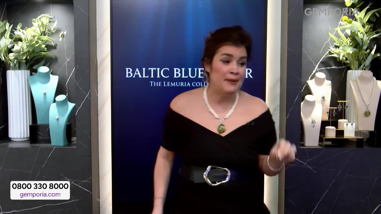A woman in a black dress gestures animatedly, showcasing a necklace with a large green pendant. Behind her, a screen displays "BALTIC BLUE LEMURIA GOLD" while jewellery pieces sit on display stands.