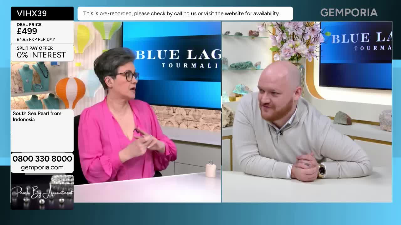 A woman in a bright pink top gestures with her hands as she speaks, while a man in a light grey jumper leans forward, listening intently. Behind them, a screen displays "Blue Lagoon Tourmaline" and a display of gemstones. A woman in a bright pink top gestures with her hands as she speaks, while a man in a light grey jumper leans forward, listening intently. Behind them, a screen displays "Blue Lagoon Tourmaline" and a display of gemstones.