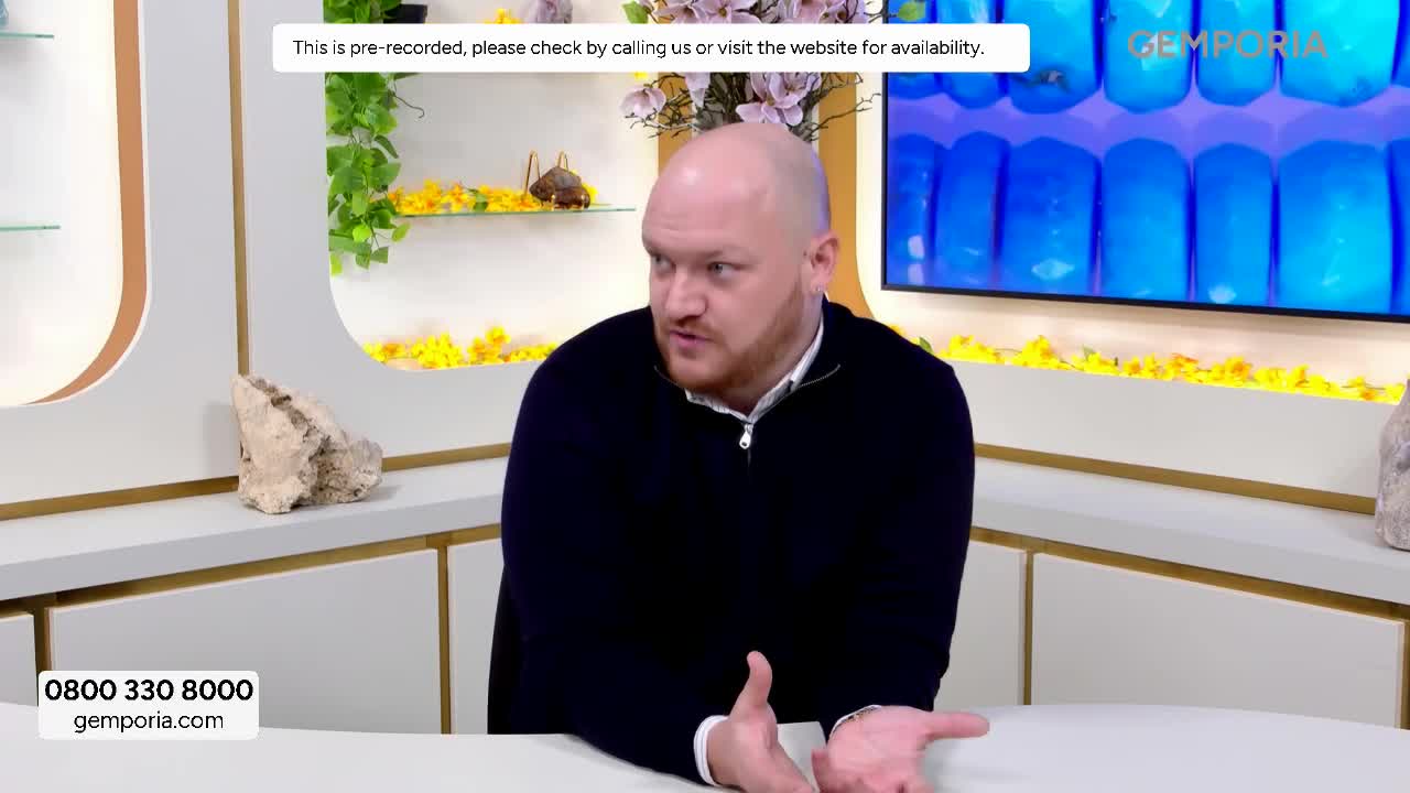 A man with a shaved head gestures with his hands while speaking. Behind him, a screen displays rows of bright blue objects, and a sign at the bottom of the screen shows a phone number and website for Gems TV. A man with a shaved head gestures with his hands while speaking. Behind him, a screen displays rows of bright blue objects, and a sign at the bottom of the screen shows a phone number and website for Gems TV.