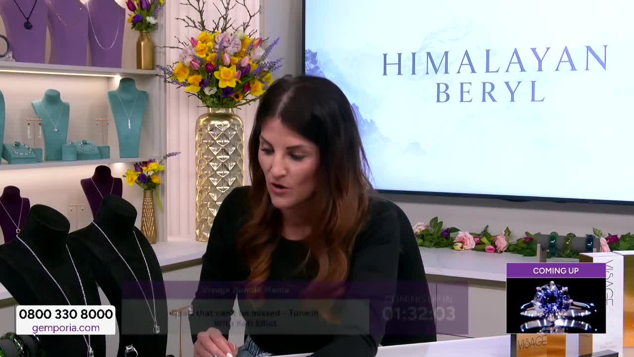 A woman is speaking in front of a display of necklaces. Behind her, a screen announces "HIMALAYAN BERYL." A woman is speaking in front of a display of necklaces. Behind her, a screen announces "HIMALAYAN BERYL."