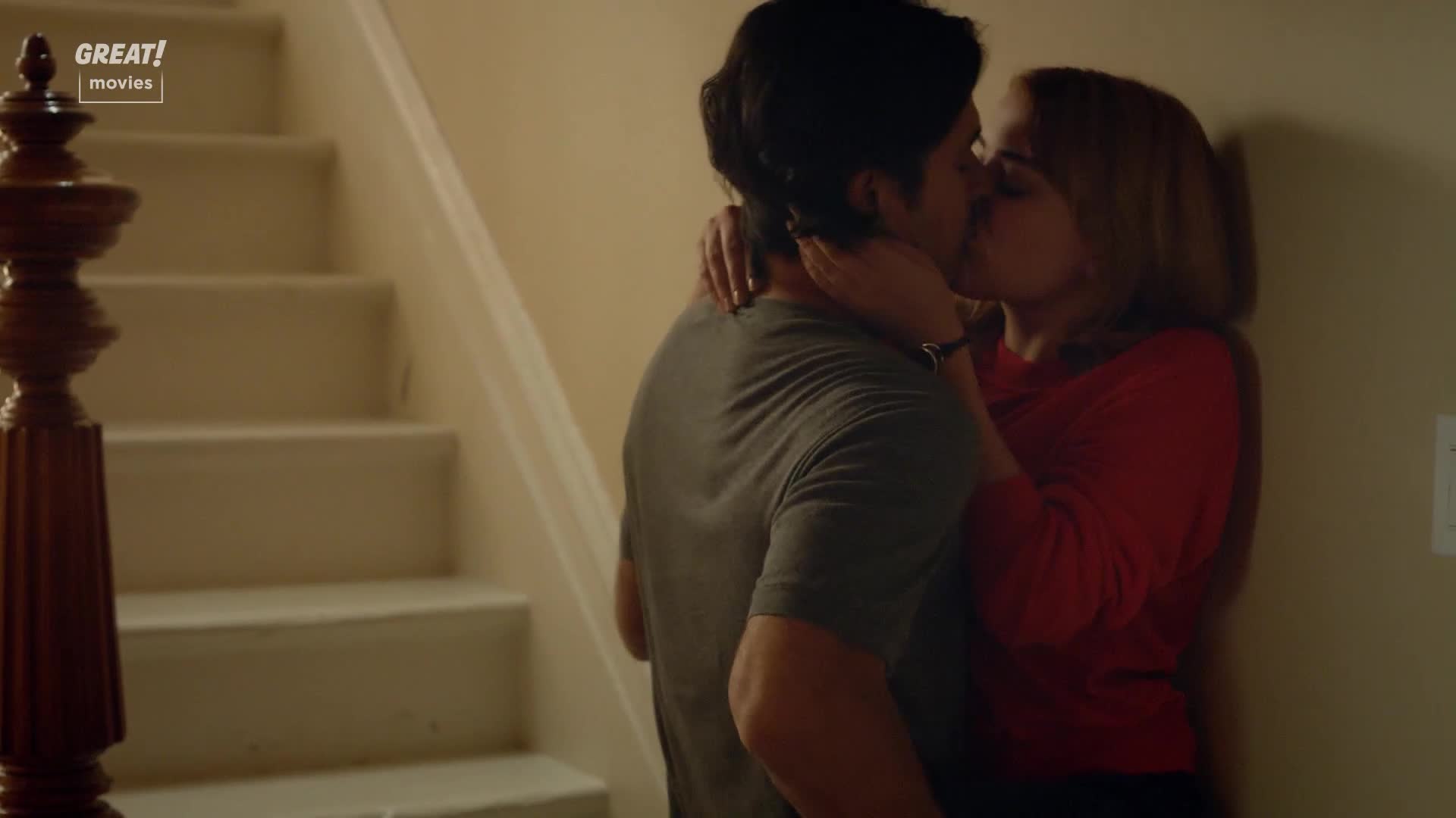 A couple is locked in a close embrace near a staircase. The woman in red presses against the wall as they kiss.
