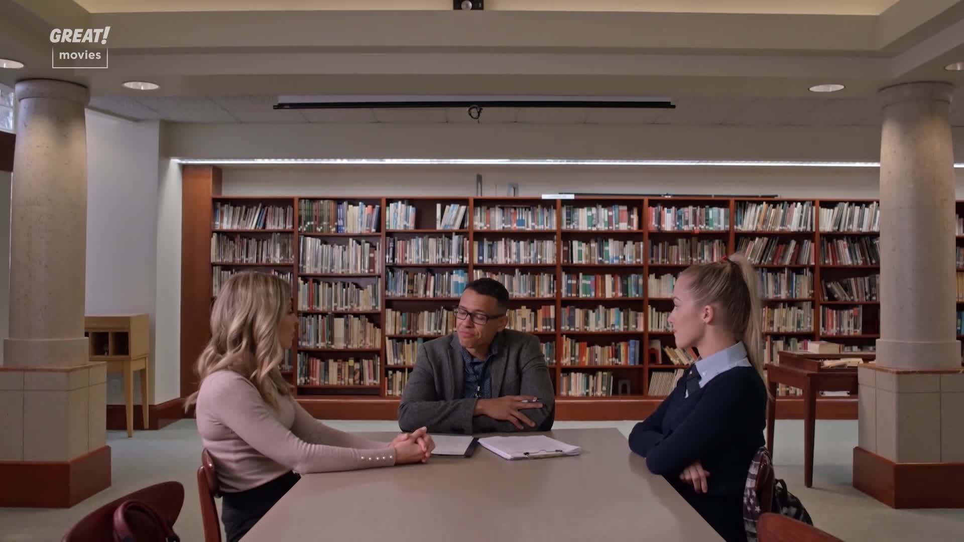 Three people sit around a table in a library, facing each other. The man in the middle is speaking, while the two women listen intently, their hands clasped in front of them.
