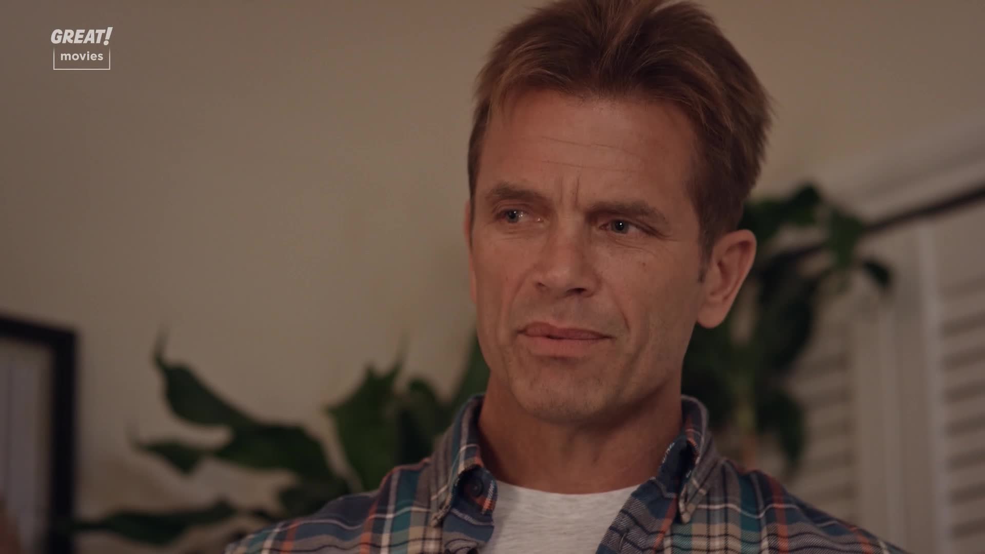 He's listening intently, his brow furrowed slightly as he considers something. The plaid shirt and the "GREAT! movies" logo suggest a cozy, made-for-TV film from the United Kingdom.
