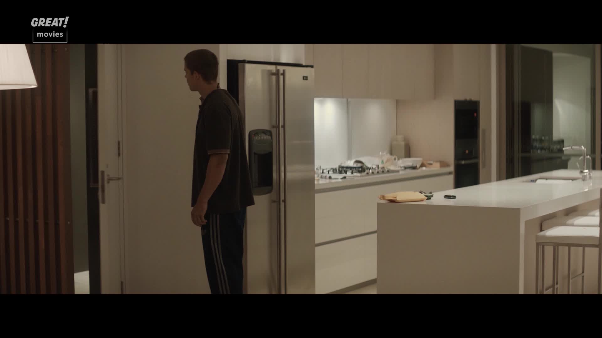 A young man in a dark shirt and track pants stands in a modern kitchen, looking toward a doorway. The space is pristine, with a large island counter and stainless steel appliances.
