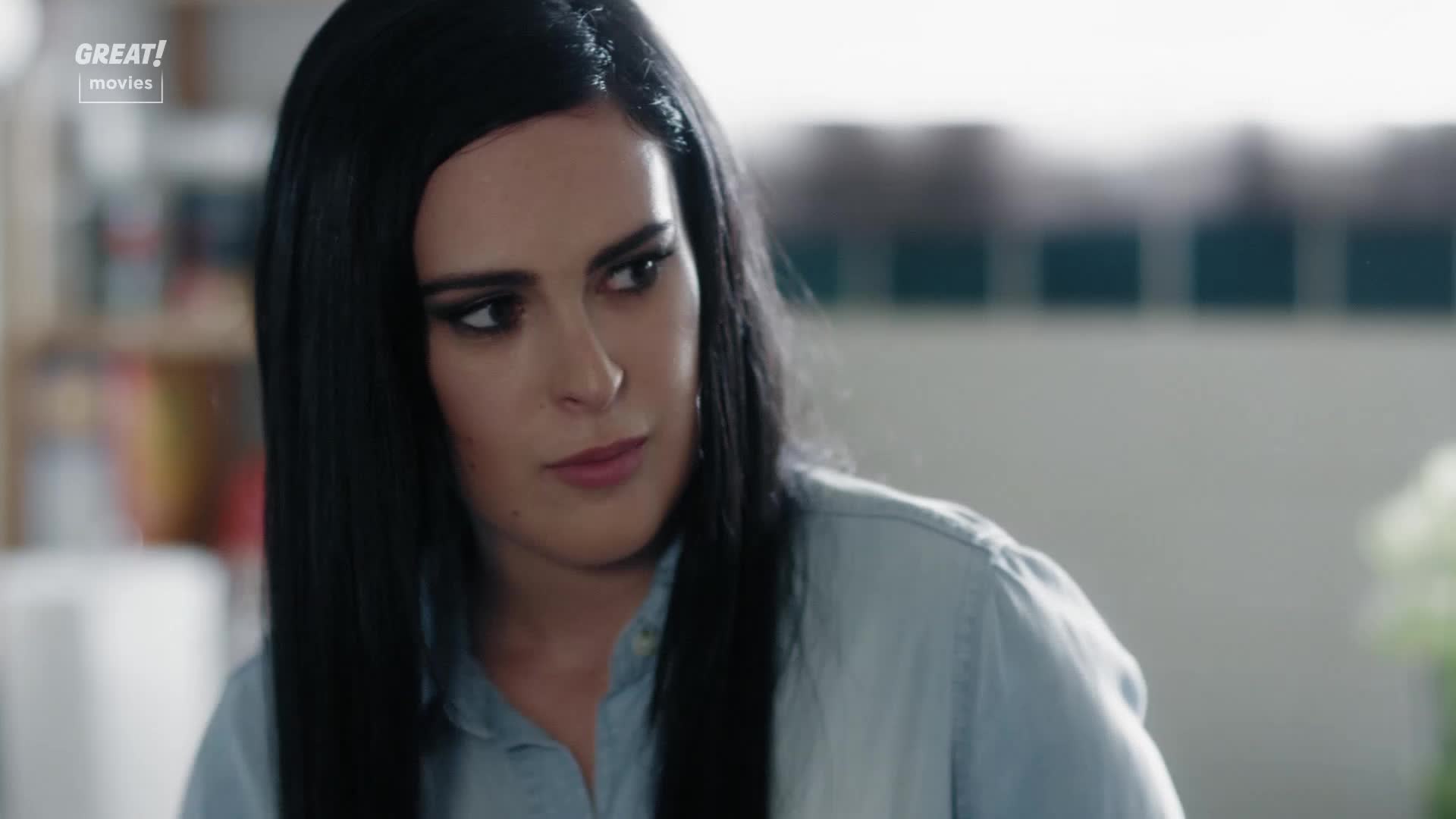 Her gaze is fixed off-screen, a slight furrow in her brow suggesting contemplation. The woman's dark hair frames her face, and she wears a light blue shirt, a scene likely from a GREAT! Movies production.
